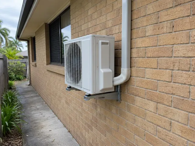 Wall-mounted outdoor air conditioner on brick exterior beside a side path in Currumbin Waters