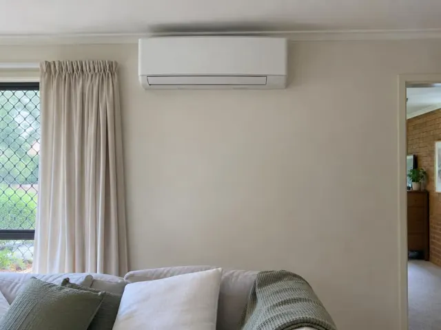 Wall-mounted split system above a sofa in a carpeted living room with curtains and a doorway in Elanora